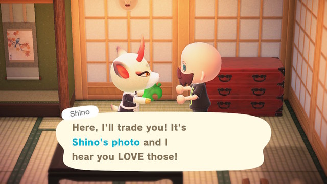 Animal Crossing: New Horizons screenshot. Set inside Shino's home which has light colored wooden screen walls, a low table in the foreground, and a reddish Japanese dresser in the background. Shino (white deer with yellow eyes and long red horns) is wearing a maid's dress and handing a green leaf to the human villager (bald, light skinned, blue eyes, bushy brownish beard, wearing a black t-shirt). Shino is saying, "Here, I'll trade you! It's Shino's photo and I hear you LOVE those!"