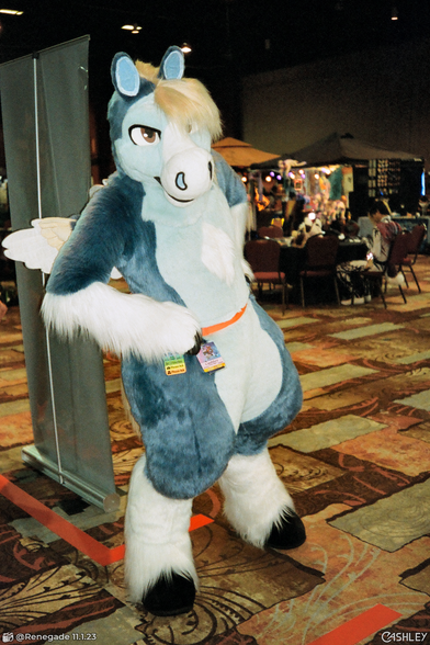 A candid film photograph of a blue pegasus fursuiter with small wings, standing in the dealer's room at BLFC 2023. He has his hands on his hips and is slightly leaning towards the camera.