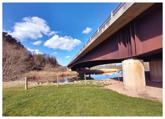 Colour photograph showing a steel and concrete modern bridge over a still river.  The sky above is blue with fluffy white clouds.