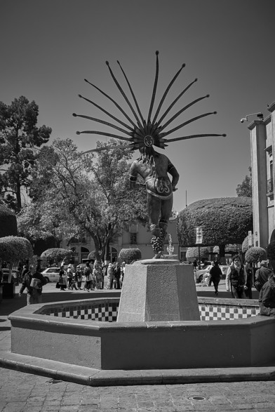 View from behind of a fountain and statue honoring the native people of central Mexico. The statue shows a male dancer in cultural garb, including a show-stopping headpiece with long branches or feathers extending for a meter or so.