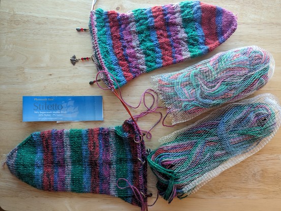 Two skeins of yarn contained in mesh netting with two socks being knit toe-up on dual circular needles (Red Lace Chiaogoo) sitting on a blond wood table.

One sock is facing up showing a pattern with chevrons and cables, the other sock is facing down showing the narrow sole.

The Red/Green/purple/teal/pink self-striping yarn with occasional streaks of primary blue has gold Lurex thread woven into the yarn for a bit of sparkle.

There are stitch markers marking the middle the sole that are split rings with a pin wrapped to it holding some black and red beads, capped with a small pewter bee. There are some smaller split ring stitch markers of pewter ladybugs marking the edge of gussets. There is also a wrapper label showing the yarn to be Plymouth Yarns Stiletto.