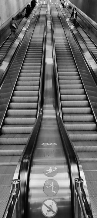 Black and white photo of an escalator in a metro station in Budapest,  Hungary. Looking up the middle of a double sided up/down escalator with people at the top travelling in both directions.