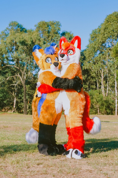 Two orange fox fursuiters in full fursuit hugging each other. The left has blue accents and the right has red accents.