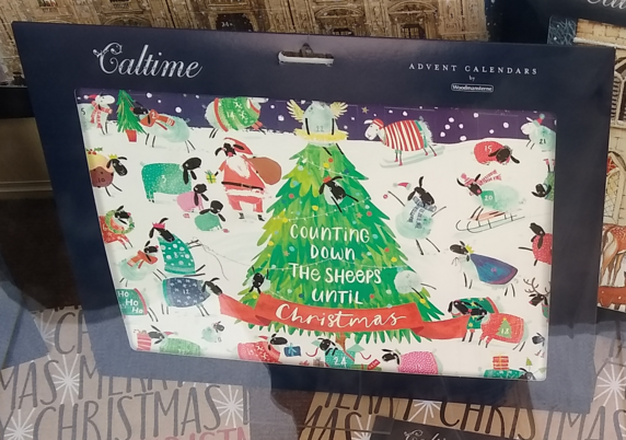Photo of a packaged card advent calendar in a shop window. The calendar's drawn snow scene is filled with images of sheep wearing jumpers or coats in various colours, one being dressed as Father Christmas. A central Christmas tree bears the words "Counting down the sheeps until Christmas".