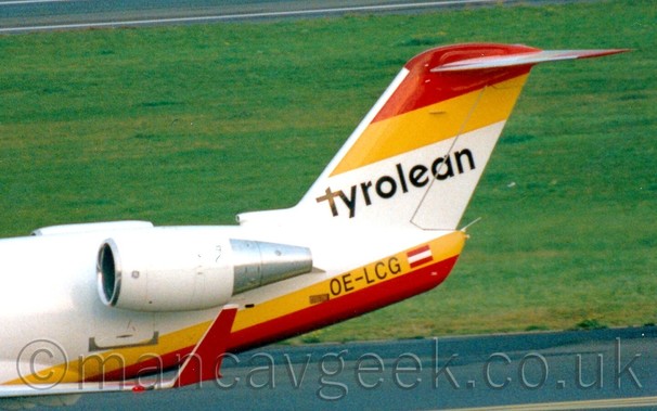 Close up of the tail of a white, twin engined jet airliner, with the engines mounted on the rear fuselage, and yellow and red stripes  at the top and bottom of the tail. with black "Tyrolean" titles in the middle.  
Grass fills the background.