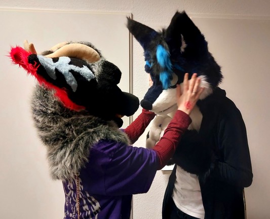 A picture of me in suit looking a bit shy, while facing another (dragon) fursuiter, who is petting my cheeks.