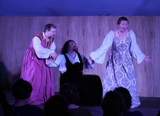 Three actors perform a commedia dell'arte play on a raised wooden stage in a pavelion tent during a medieval camping event portraying three young lovers in dresses.