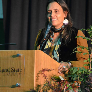 A photo of Winona LaDuke behind a wooden podium that says "-land State." She is smiling at the audience. She is holding the side of the podium, which has a plant next to it. She has medium brown skin and long dark hair. She is wearing an orange shirt, black vest, blue and green scarf, and large circular abalone earrings.