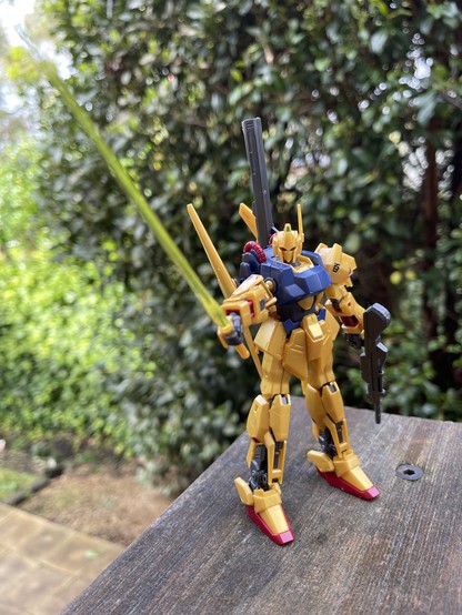 A wide colour photo of a small toy robot, gold and blue. It's holding out a yellow energy sword. In the background: grass and tree foliage.