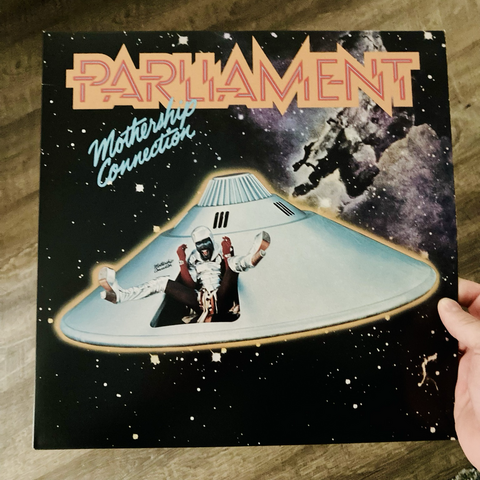Album cover for Mothership Connection by Parliament, featuring a photo of a dome-shaped spaceship in space, a wild spaceman with legs outstretched hanging out of a bay door.