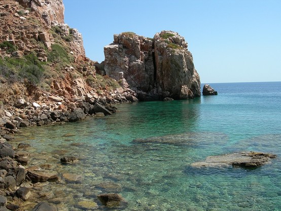 a photo of the beach, some cliffs coming out of the water, the sky is sunny and blue, the water clear, you can see the rocks under the water easily
