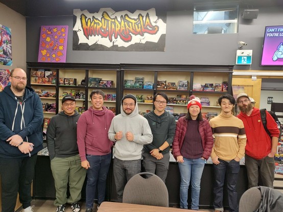The top 8 players in the Lorcana Rise of the Floodborn release event stand together in front of a sign that says "Hairy Tarantula"