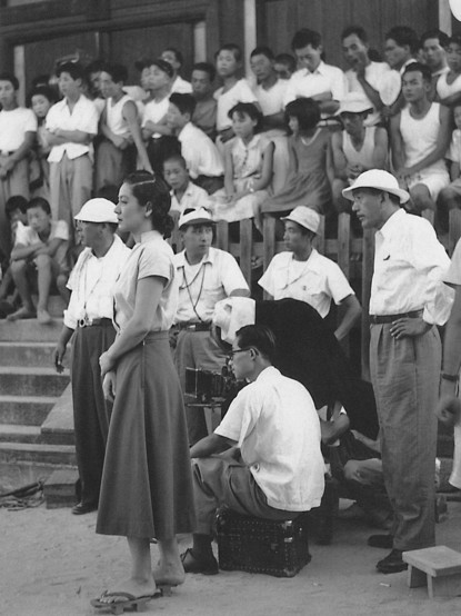 A group of people are gathered outdoors. Some sit down, other stand. The director oversees the cameraman's work. The actress is waiting for her turn to join the scene.