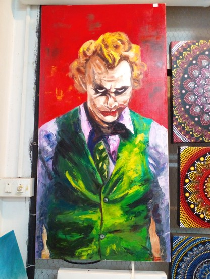 a painting of the Joker, a red background, he is seen from his waist up, his arms at his sides, he is wearing a purple dress shirt, green vest, and green tie, he looks at the viewer with a menacing smile