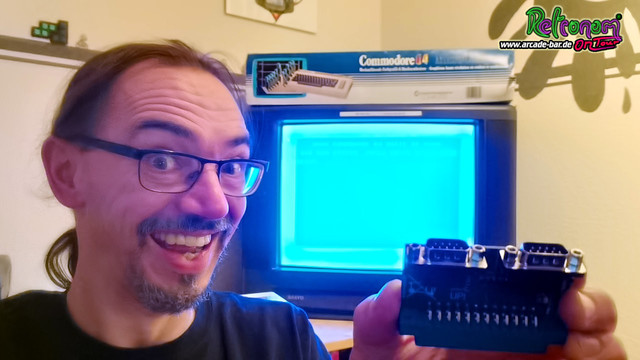 Mattus hält den Vier-Spieler-Adapter hoch. Im Hintergrund ist ein Röhrenfernseher zu sehen, auf dem der C64-Startbildschirm gezeigt wird und obenauf liegt ein Commodore 64-Karton.