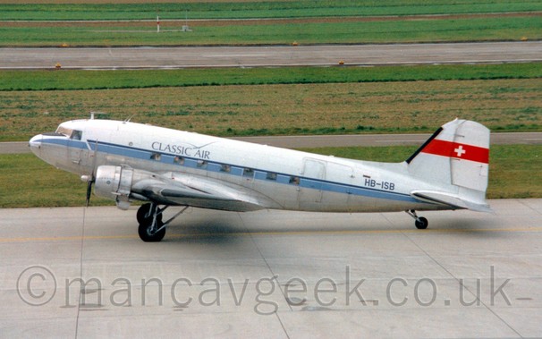 Side view of a white and grey twin propellor engined airliner taxiing from right to left. Green grass and grey taxiways fill the rest of the frame.