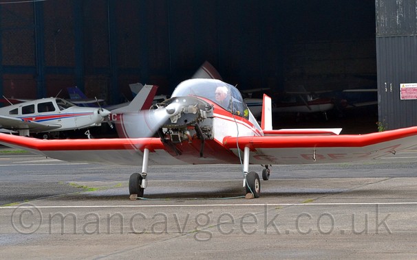 Front view of a red and white, single englined light aircraft, with it's engine cowling missing, performing an engine test run, parked facing almost directly towards the camera. In the background is a black hangar, filled with more light aircraft.