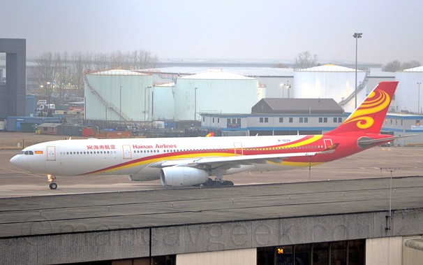 Side view of a white, twin engined jet airliner with a red and yellow stripe running along the body before sweeping up in to the tail, with red "Hainan Airlines" in both Chinese and English on the upper forward fuselage, taxiing from right to left, under a hazy grey sky.