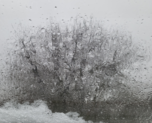 Regentropfen und matschiger Schnee auf einer Fensterscheibe. Im Hintergrund ist unscharf eine Baumkrone ohne Blätter zu erkennen. Der Himmel ist grau.