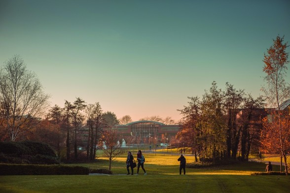 A photograph taken on the University of Limerick campus. It shows a vast law stretching into the distance with many groups of trees. In the foreground a group of students are walking across a path on the lawn, and in the distance is a tall glass-fronted building. The sun is low in the sky on what looks to be a cold day