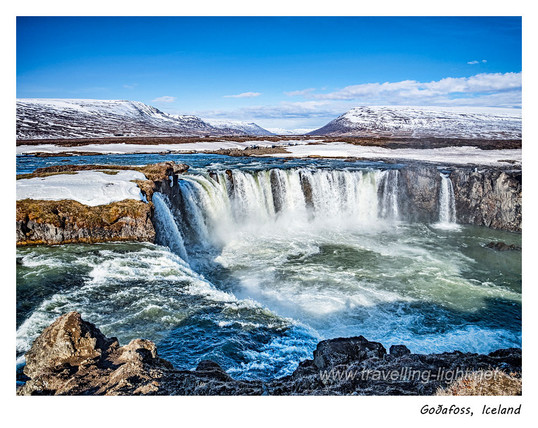 A wide horseshoe shaped waterfall in a landscape with some snow cover, mountains in the distance, and a blue sky.