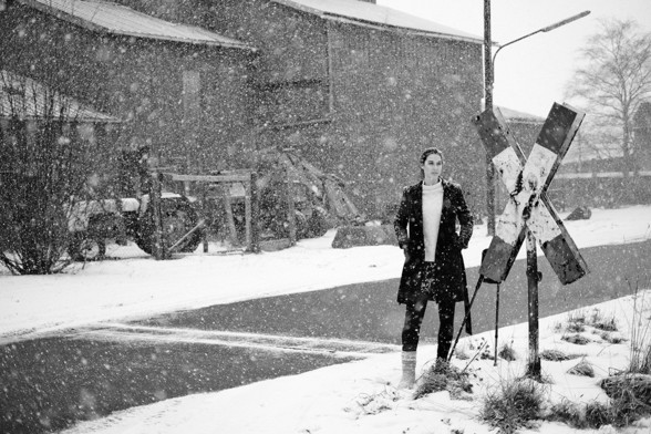 Junge Frau steht am Straßenrand bei heftigem Schneefall. Ihre Krücken sind an ein Andreaskreuz gelehnt. Ihr rechts Bein steckt in einem Gips, am Fuß trägt sie eine dicke Socke.