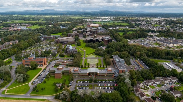 An aerial photograph of the University of Limerick campus. It shows many orange and brown brick buildings nestled amongst dense mature tress, connected by a series of lawns