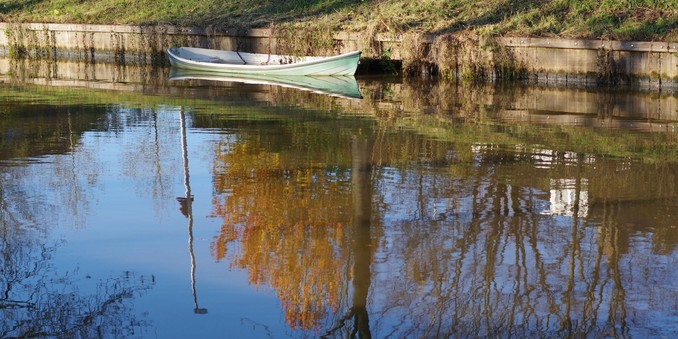 Photograph of a canal with still water, with a small green-gray painted boat moored on the shore visible. The boat is reflected in the surface of the water, along with trees in fall foliage.