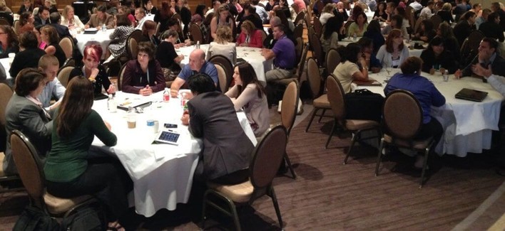 improve your facilitation: a photograph of a Solution Room conference session, with participants talking animatedly in groups of eight at round tables