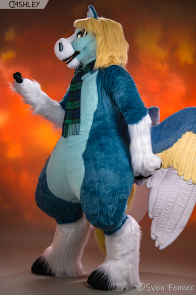 A full-body studio portrait of a blue pegasus fursuiter with big wings, standing in front of an orange autumnal blowing-leaf backdrop. He's wearing a dark blue and green tartan scarf and turned to face camera-left, with his right arm raised and his left arm relaxed.