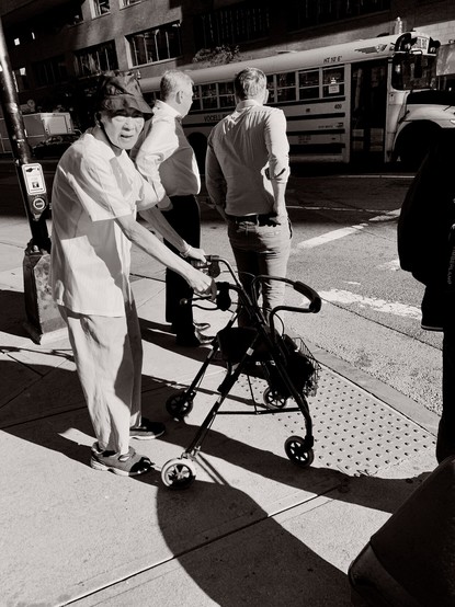 A black and white photo of a person standing on a street corner with a walker.