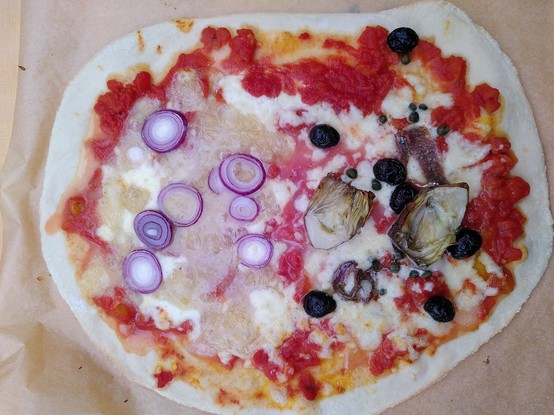 Pizza auf einem Holzbrett. Zwei verschiedene Beläge: eine Seite Graukas mit Zwiebelringen, andere Seite: Artischocken, Oliven, Sardellen und Kapern
