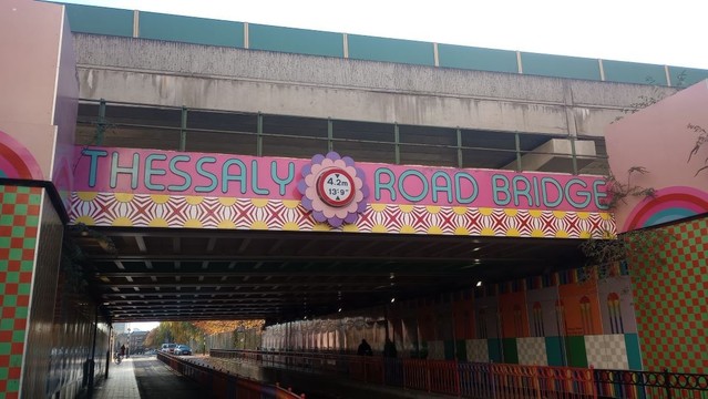 A low, concrete bridge looms over a gloomy stretch of road. The bridge is a two-layered affair, carrying railway lines at two different levels (eight in total, I think).
The upper deck is concrete with a functional green fence across the top of it. The lower deck has a green-painted fence, but the concret has been painted mainly pink, with a lower geometric margin of in red, gold, white and blue.
Across the pink portion is written "Thessaly Road Bridge" in large turquoise letters. Between the first two words, in the centre of the bridge, is a large rosette of purple and yellow. In the centre of the rosette is a white, round, red-bordered warning sign declaring 4.2m and 13' 9".