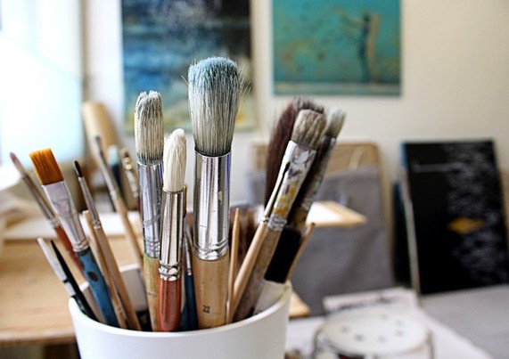 Paint brushes in a cup in an artists' studio with out of focus paintings in the background
