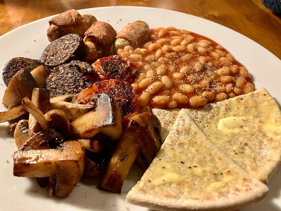 Modified vegan English breakfast plate featuring potato farls, mushrooms, black pudding slices, pigs in blankets, baked beans and black pepper