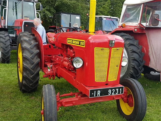 A vintage David Brown tractor. The tractor is red with yellow details, such as the front grille, exhaust and wheel centres.
The tractor fills most of the frame, and is parked with other classic tractors at a show.