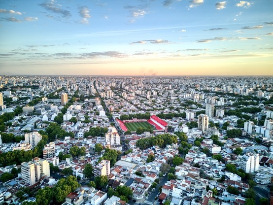 Sunset drone image of a huge metropolis spreading till the horizon. In the middle of the built area a soccer stadium occupies an entire block. Tribunes are painted in red.