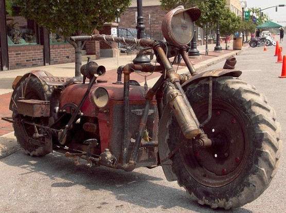 A "rat bike", crudely assembled motorcycle, though this one has a lot of ingenuity in that it uses large diameter tractor wheels and tires, and the tractor engine cowl has been preserved over the engine in its normal place between the wheels. A tractor seat (natch) completes the build.