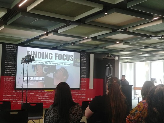 Filmmaker Dan Sanguineti talking at a lectern in front of an audience, near a large screen with a still picture of him holding a camera and the headline 'Finding Focus: Unmasking my Filmmaker's Story'