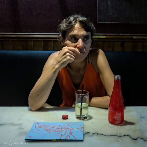 A woman sitting on a bench in a bar in front of a glass of a soft drink with lemon. She looks to the right. Elements in red tones: the dress, the bottle and its capsule, the streets on a map.