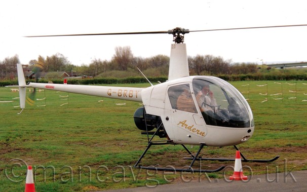 Side view of a white light helicopter, parked facing to the right, on grass, with more grass and trees in the background under a grey sky.