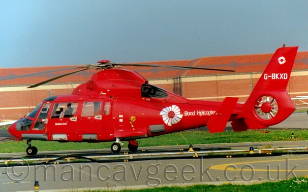 Side view of a red helicopter parked facing to the left, with a red brick building filling the background, under a blue sky.