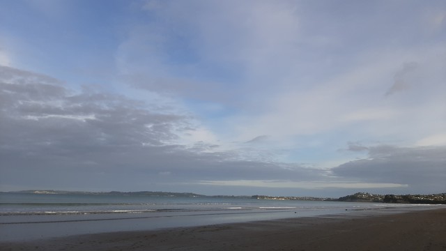 A beach at low tide. A pallette ofblight blues and greys. A long bay extends from left to right finishing in a low headland. Large sky with broad but light clouds.