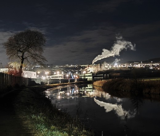 A mainly dark early morning picture with the canal and towpath  leading into the darkness of an industrial estate. a tree silhouette frames the left of the photo. The bright lights of the buildings light up the industrial units and a plume of smoke or steam is reflected on the surface of the water.