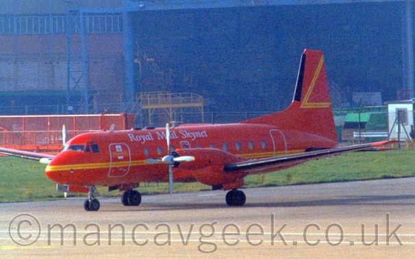 Side view of a red, twin propellor-engined airliner  with a yellow cheatline running along the body and white "Royal Mail Skynet" titles on the upper forward fuselage, parked facing to the left, with a large black hangar in the background.