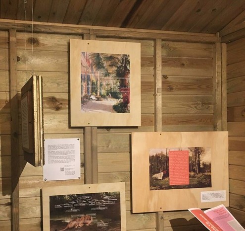 Corner of a wooden hut, with an exhibition of three printed paintings. Of a palm house, a lion, and an outdoor scene in a forest. On top of the two paintings on the left are diagrams of text, on the right a red block and white text. Below the painting on the right hangs a little magazine.
