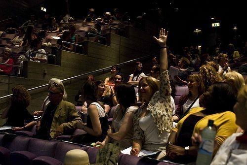 minimize vendor pitches: photograph of a conference audience with one attendee raising their hand to ask a question