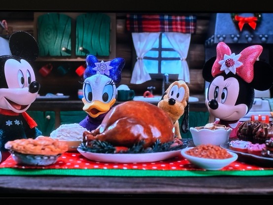 Mickey Mouse, Daisy Duck, Pluto, and Minnie Mouse all sit around a holiday table featuring a large roast turkey in the middle.