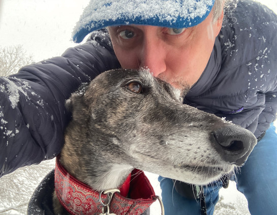 Selfie of me kissing the top of Taylor the greyhoundâ€™s head, my jacket and hat brim dotted with snowflakes, as are the dogâ€™s snout and eyelashes.