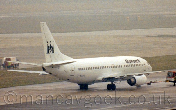 Side view of a an all-white twin engined jet airliner with black "Monarch" titles on the upper forward fuselage and a black capital "M" shaped like a crown on the tail, facing to the right and slightly away from the camera, being pushed back from it's stand by a red and white tug attached to the nosewheel.
In the background, taxiways and grass rapidly vanish in to mist.